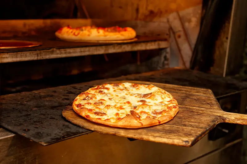 Close-up of cheesy pizza on a wooden peel being baked in a hot oven