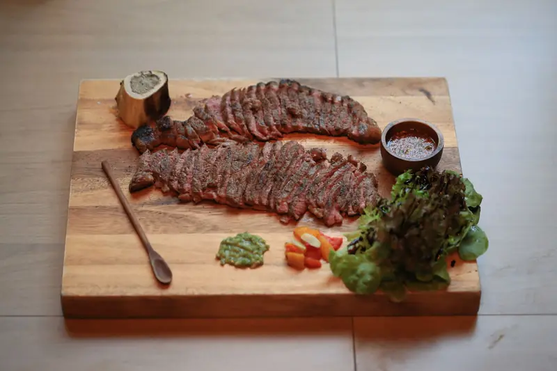 Sliced grilled steak with green pesto, sauce, and lettuce on a wooden cutting board