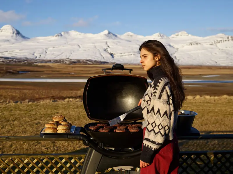 Side view of thoughtful young female traveler in warm clothes looking away and grilling steak during picnic in countryside in winter