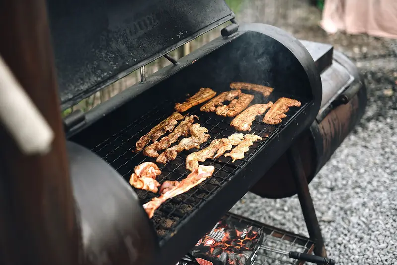 Close-up of a barbecue grill outdoors with various meat strips cooking, exuding rich smoke