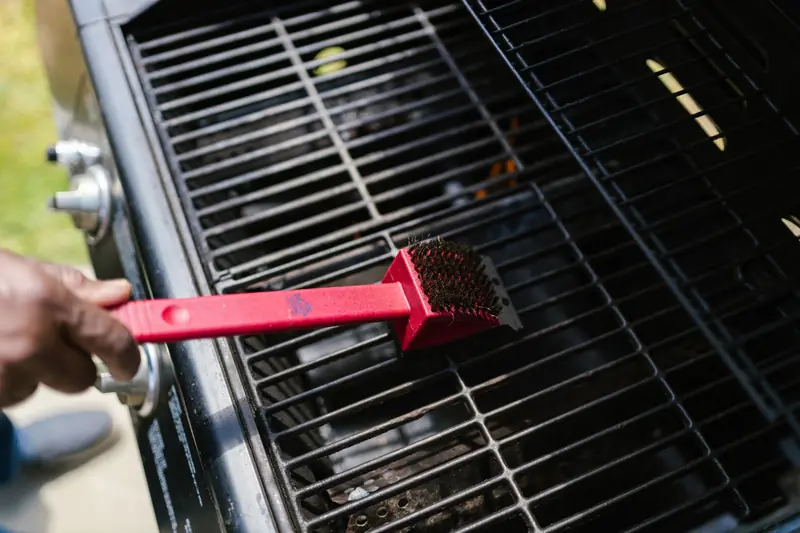 A person cleaning a barbecue grill with a red brush in daylight. Ideal for BBQ enthusiasts